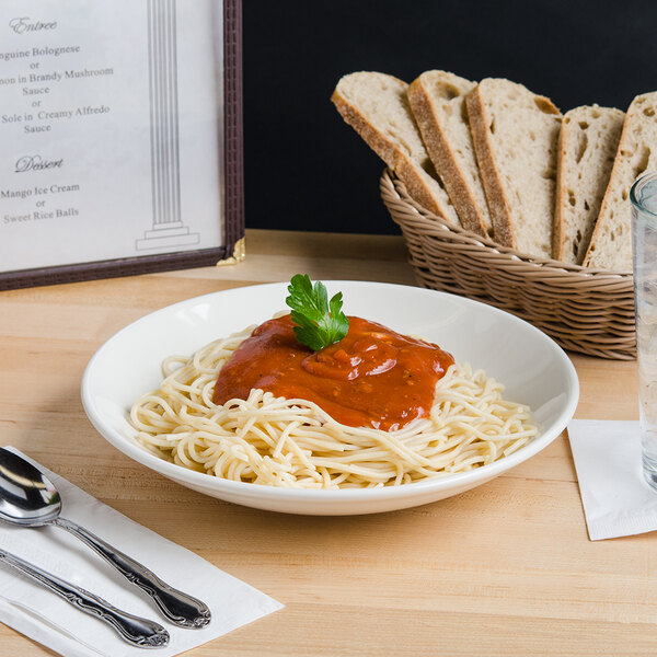 A Tuxton eggshell china bowl filled with spaghetti with sauce and parsley on a table with a fork and a glass of water.