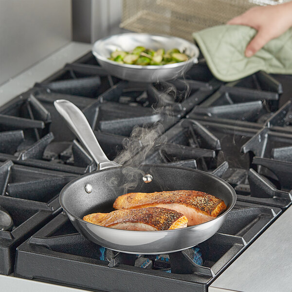 A hand using a pot holder to cook food in a Vollrath stainless steel non-stick fry pan.