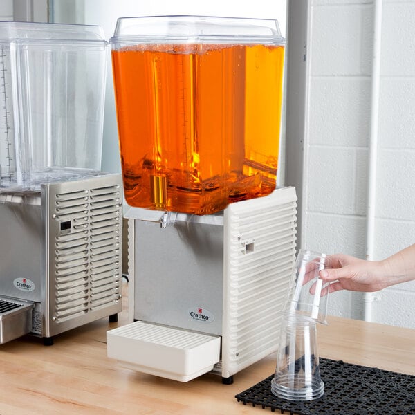 A person using a Crathco refrigerated beverage dispenser to pour orange juice into a plastic cup.