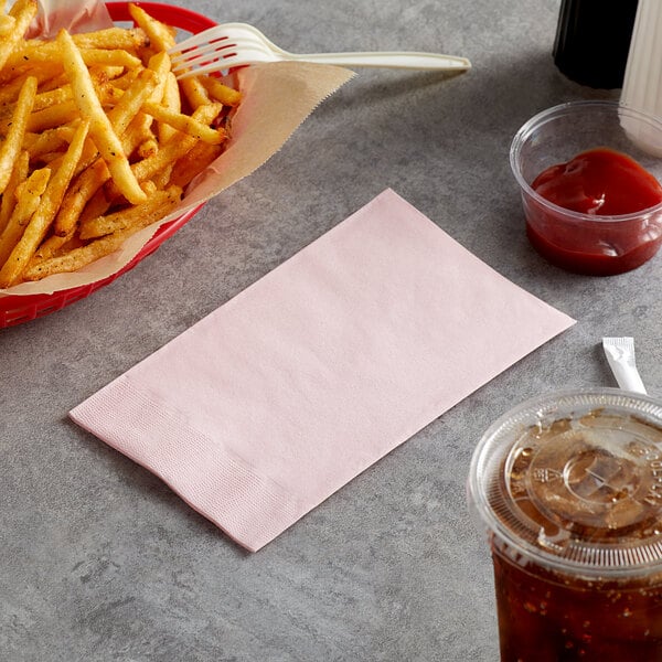 A basket of french fries next to a pink paper dinner napkin and a drink.