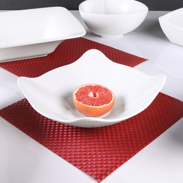A white Elite Global Solutions square melamine bowl filled with a grapefruit half on a red surface.
