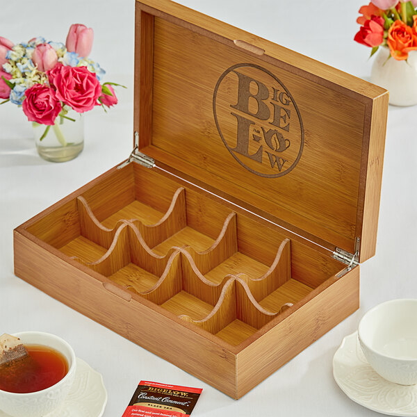 A wooden Bigelow Tea chest open with tea and a cup of tea on a table with flowers.