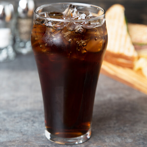 A Libbey customizable bell soda glass filled with brown soda and ice on a table with a sandwich.
