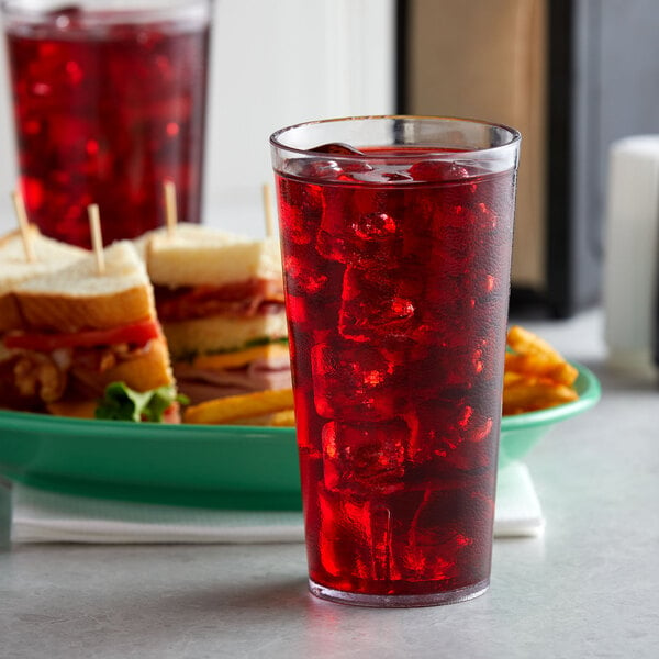 A close-up of a clear plastic tumbler filled with red liquid and ice on a table with sandwiches.