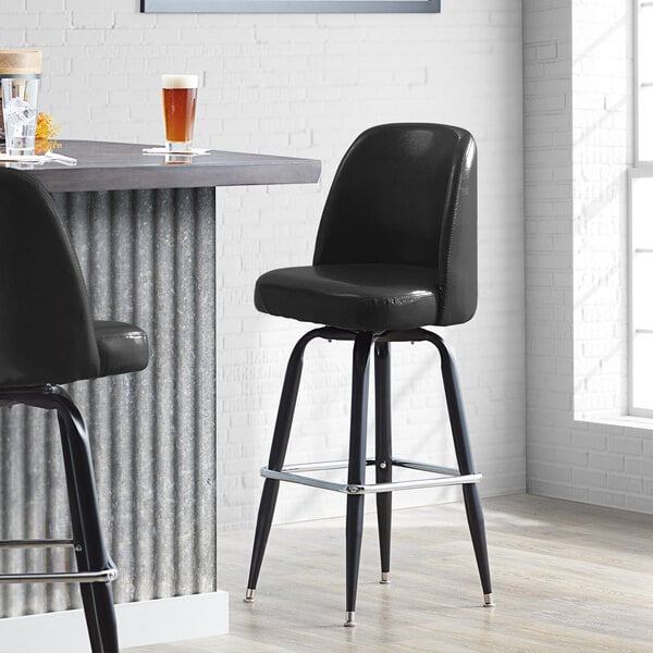 A black Lancaster Table & Seating bar stool with metal legs next to a table with a beer glass.