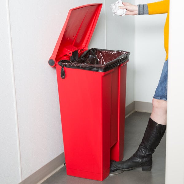 A woman in a yellow shirt and boots standing next to a red Rubbermaid commercial trash can.