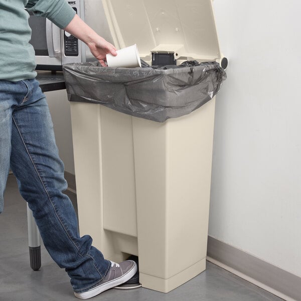 A person in blue jeans using a Rubbermaid beige rectangular mobile step-on trash can.