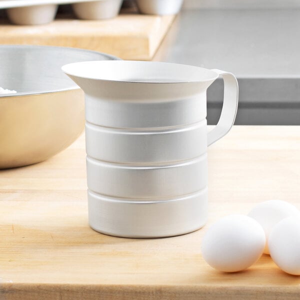 A white Vollrath aluminum measuring cup on a wooden table next to eggs.