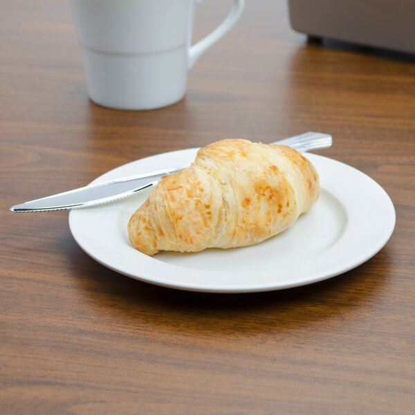 A Tuxton AlumaTux white china plate with a croissant on it on a table with a white mug and knife.
