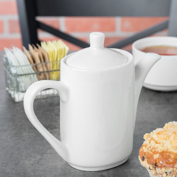 A white Tuxton china coffee pot on a table with a cup of tea.