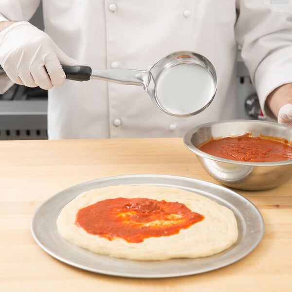 A person in a white coat using a Vollrath black solid round Spoodle to pour red sauce onto a pizza.
