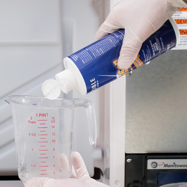 A gloved hand pours blue liquid from a Noble Chemical QuikSan can into a measuring cup on a counter.