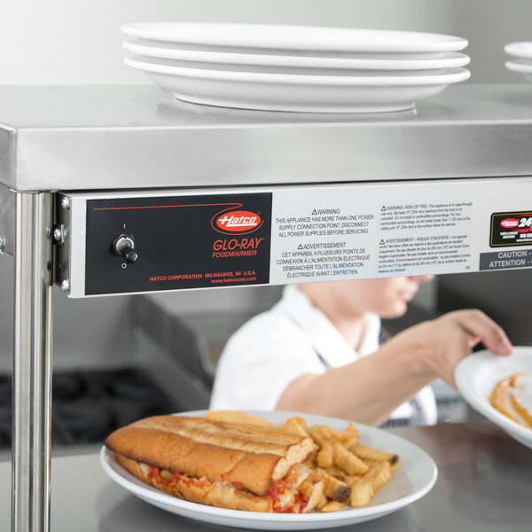 A Hatco dual infrared warmer above plates of hot dogs, fries, and a sandwich on a counter.