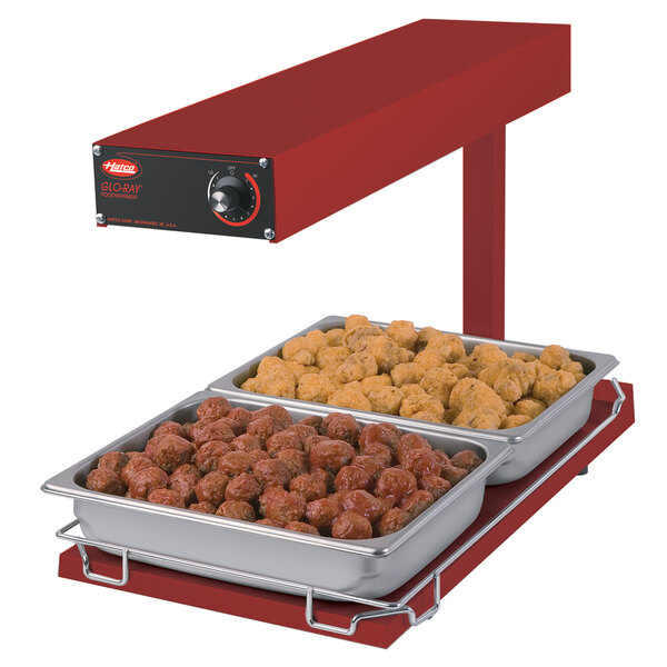 A red Hatco Glo-Ray heated shelf food warmer on a table with trays of meatballs and chicken.