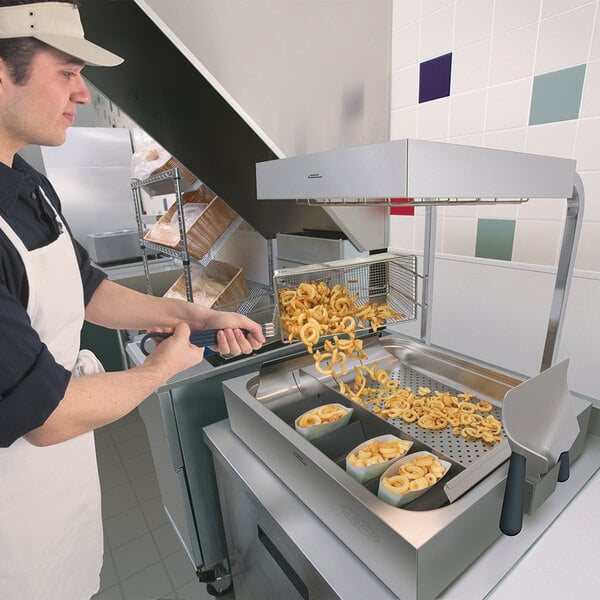 A man in a white apron using a Hatco fry holding station to prepare french fries and onion rings.