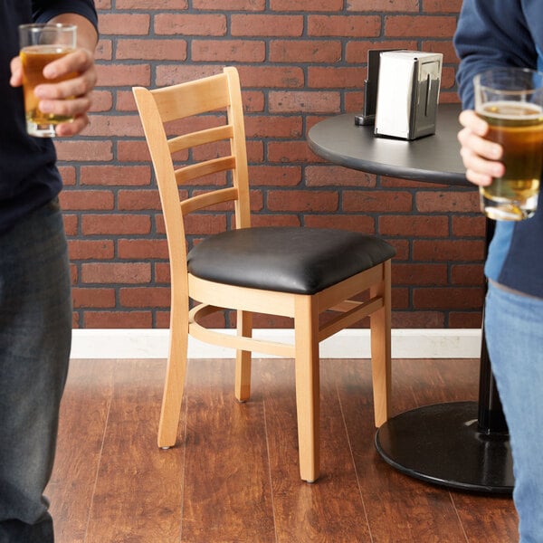 A Lancaster Table & Seating wood ladder back chair with a black vinyl seat next to a table with glasses of beer.