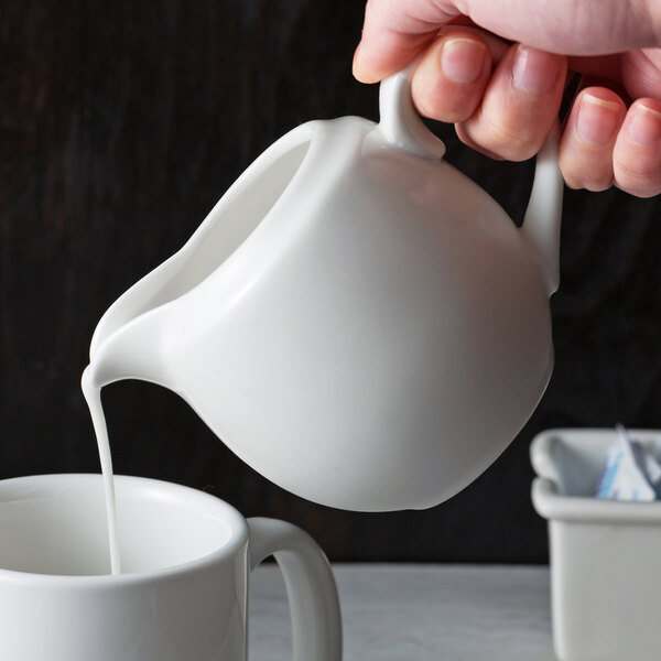 A person pouring milk from a white 10 Strawberry Street Whittier creamer into a white cup.