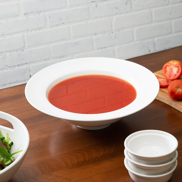 A white 10 Strawberry Street Whittier rim soup bowl filled with red soup on a table with a salad bowl.