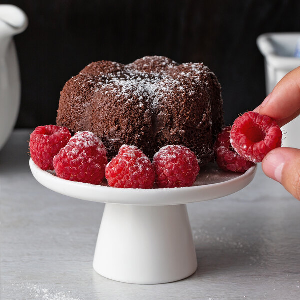 A hand holding a raspberry over a chocolate cake on a white cake stand.