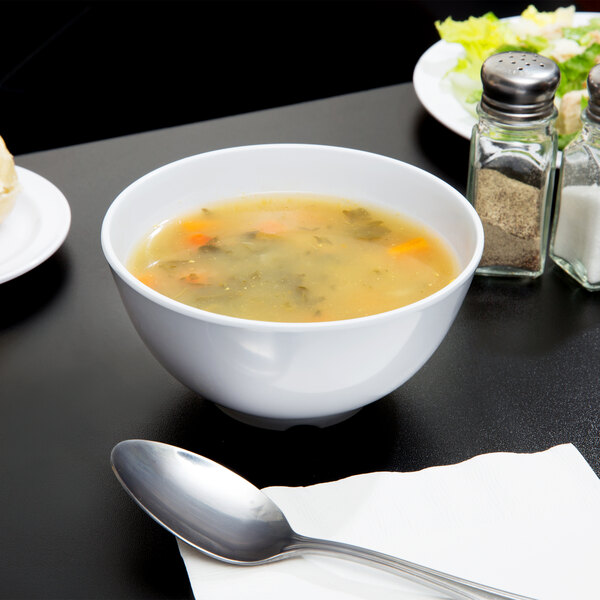 A white melamine bowl of soup on a table with a silver spoon.