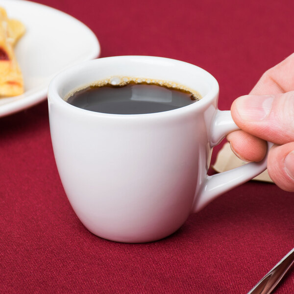 A hand holding a Tuxton bright white espresso cup filled with coffee on a saucer on a table.