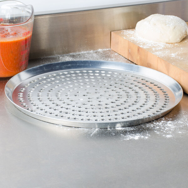 An American Metalcraft heavy weight aluminum pizza pan with perforations on a counter next to a bowl of dough.