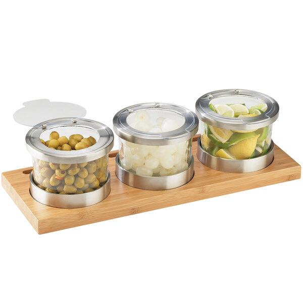 A Cal-Mil bamboo display with three glass jars and hinged lids filled with green olives, onions, and round white objects.