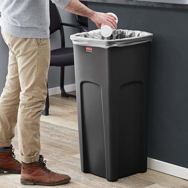 A man standing next to a Rubbermaid black rectangular trash can.