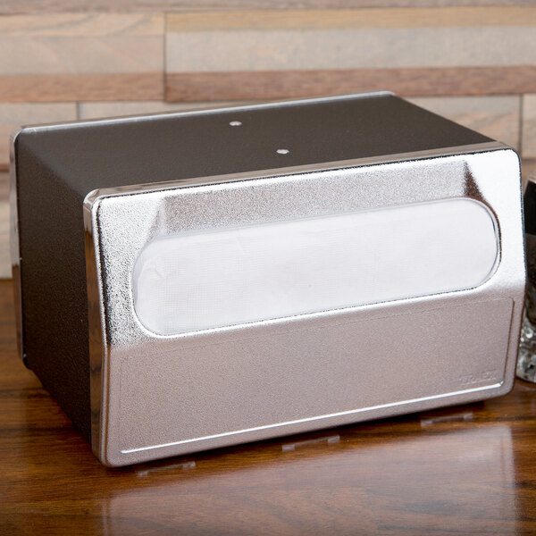 A silver metal Vollrath tabletop napkin dispenser with a clear window on a wood surface.