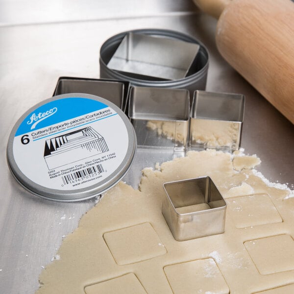 A box of Ateco stainless steel square cookie cutters on a table with dough and cookies.