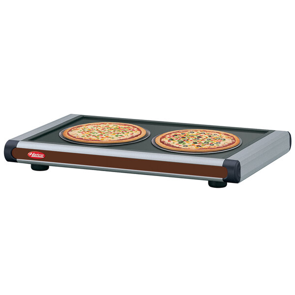 Two pizzas on a Hatco heated shelf with black caps on a table.