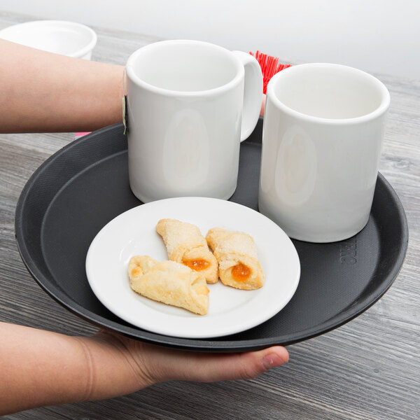 A person's hand holding a Cambro black non-skid serving tray with a plate of cookies and two white mugs.
