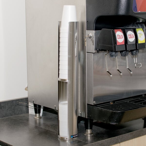 A San Jamar two-tier stainless steel lid holder on a wall above a soda machine.