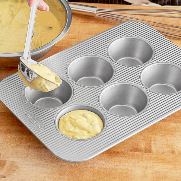 A person using a silver bowl to pour batter into a Chicago Metallic Texas Jumbo Muffin Pan.