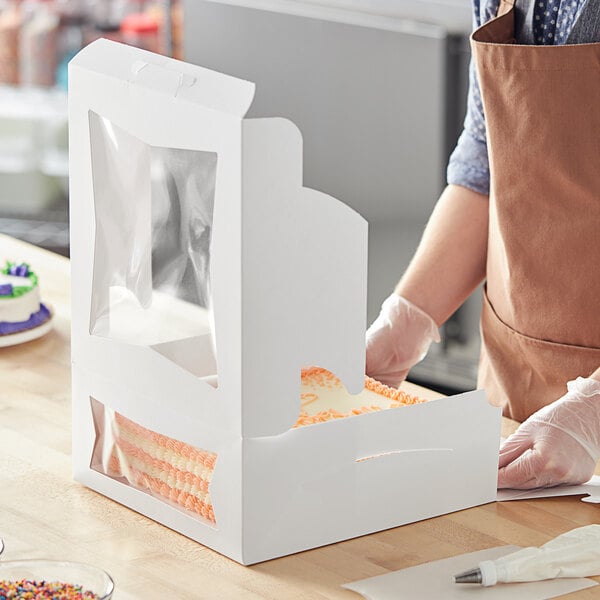 A person wearing gloves and a brown apron cutting a cake in a white Baker's Mark box with orange frosting.