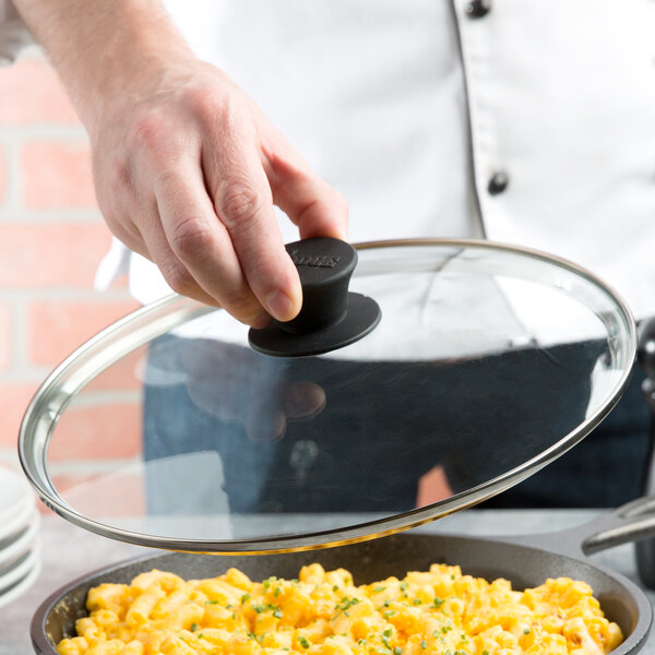 A hand using a Lodge tempered glass cover to cook food in a skillet.