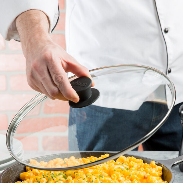 A hand holding a Lodge tempered glass lid over a pan of macaroni and cheese.