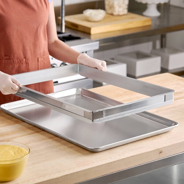 A person wearing gloves is holding a rectangular metal frame above a large metal baking sheet on a wooden countertop.
