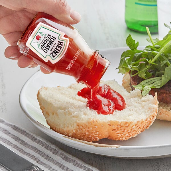 A hand pouring Heinz Ketchup from a green glass bottle onto a bun.