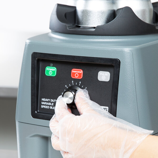 A person wearing plastic gloves presses a knob on a Waring blender control panel.
