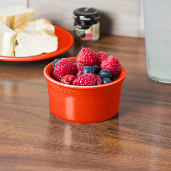 A Fiesta Poppy china ramekin filled with raspberries and blueberries sits on a table next to a plate of cheese.