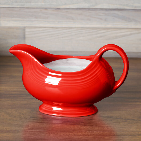 A Fiesta poppy red china gravy boat with a handle on a wood surface.