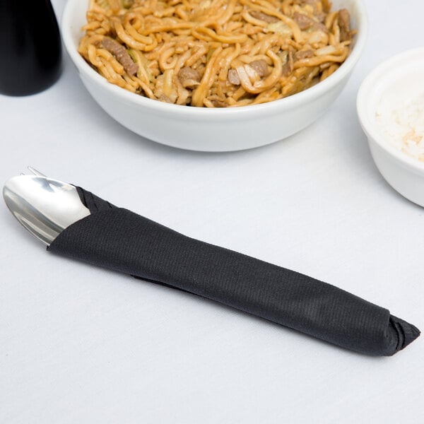 A black Flat Pack Linen-Like napkin on a table with a spoon and a bowl of noodles.