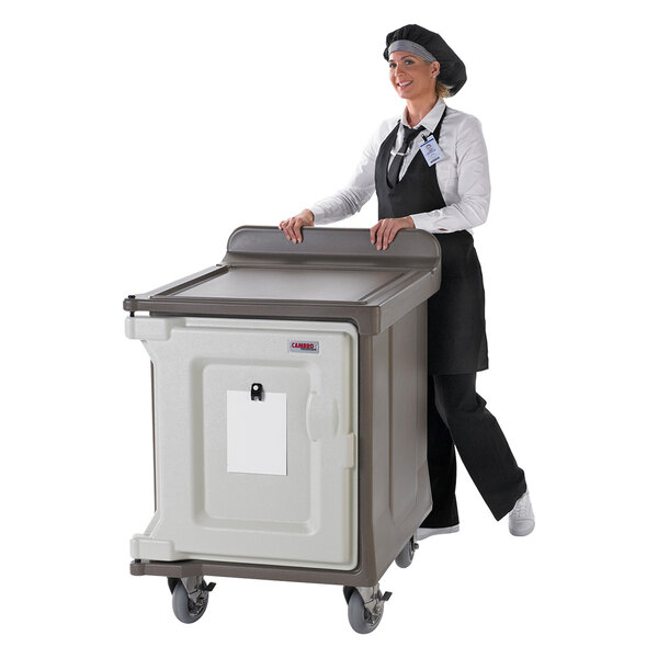 A woman in a black apron and chef hat pushing a Cambro meal delivery cart.