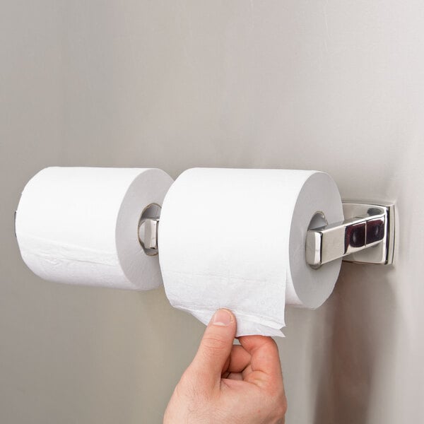 A person holding a roll of toilet paper in front of a Bobrick surface-mounted toilet tissue dispenser.