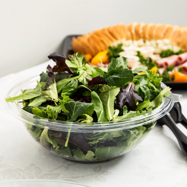 A Fineline clear plastic bowl filled with salad and a plate of food on a table.