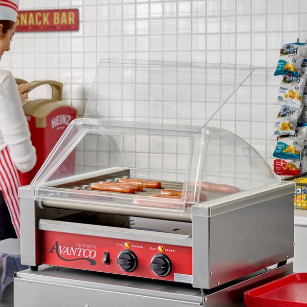 A countertop hot dog roller grill with a clear sneeze guard, containing several hot dogs inside.