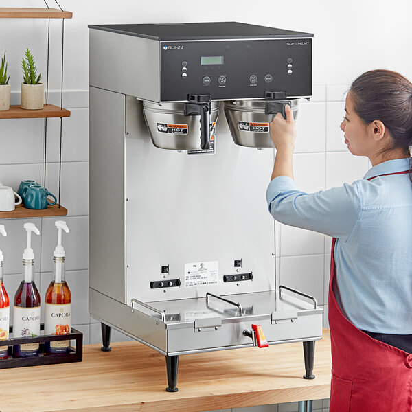 A woman in an apron using a Bunn BrewWISE dual soft heat coffee machine on a counter.