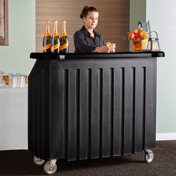 A woman standing behind a black Cambro portable bar with orange bottles on the counter.