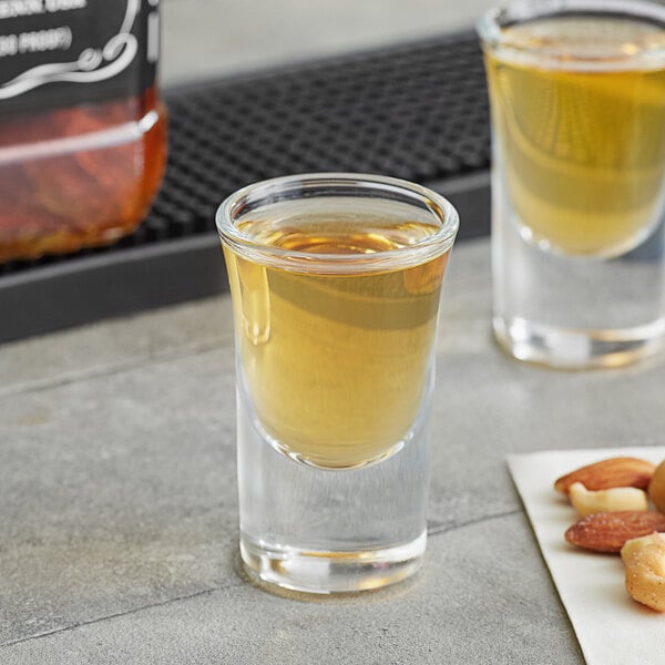 Two Acopa shot glasses filled with yellow liquid on a table with nuts.
